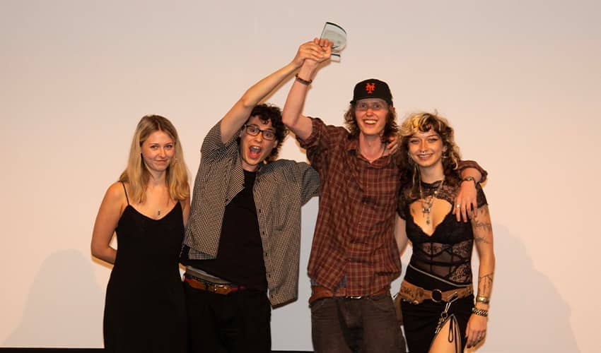 Four students stand together on a stage, smiling and celebrating, with two raising a glass award overhead. 