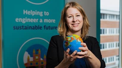 A head shot of a woman with blonde hair wearing a blue dress holding a small globe standing in front of a branded board saying 'Building a more sustainable future'