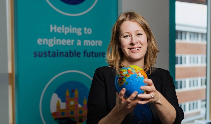 A head shot of a woman with blonde hair wearing a blue dress holding a small globe standing in front of a branded board saying 'Building a more sustainable future'