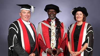 Two men and a woman who are all wearing university graduation gowns smile against a grey background.