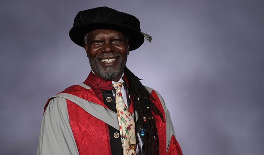 A man wearing university graduation gowns smiles against a grey background.
