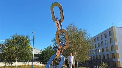 A man resting against a large stainless steel sculpture of multiple links connecting outside in an open park area