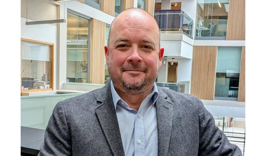 A headshot of a man wearing a suit jacket and shirt standing in a coporate building with large glass windows and desks visible in the surrounding rooms. 