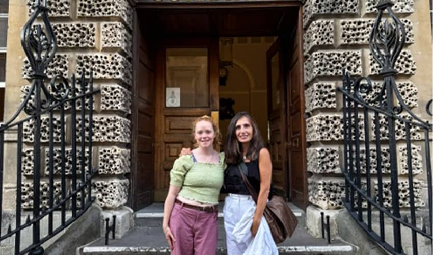 Two people stand on stone steps outside the entrance of a historic building. The doorway is framed by decorative stonework and black wrought-iron railings, with lantern-style lights mounted on either side. One person is wearing a light green top and mauve trousers, while the other is wearing a dark top and flowing white trousers.