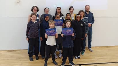 A group of school children holding up a blue book along with a group of adults standing posed towards the camera in a school sports hall setting. 