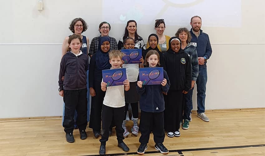 A group of school children holding up a blue book along with a group of adults standing posed towards the camera in a school sports hall setting. 