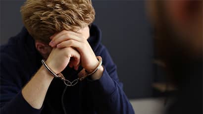 A young man wearing a hoodie with his head in his heads with handcuffs around his wrist. He is sat opposite someone who is blurred in the image.