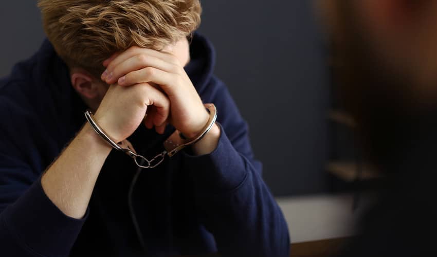 A young man wearing a hoodie with his head in his heads with handcuffs around his wrist. He is sat opposite someone who is blurred in the image.
