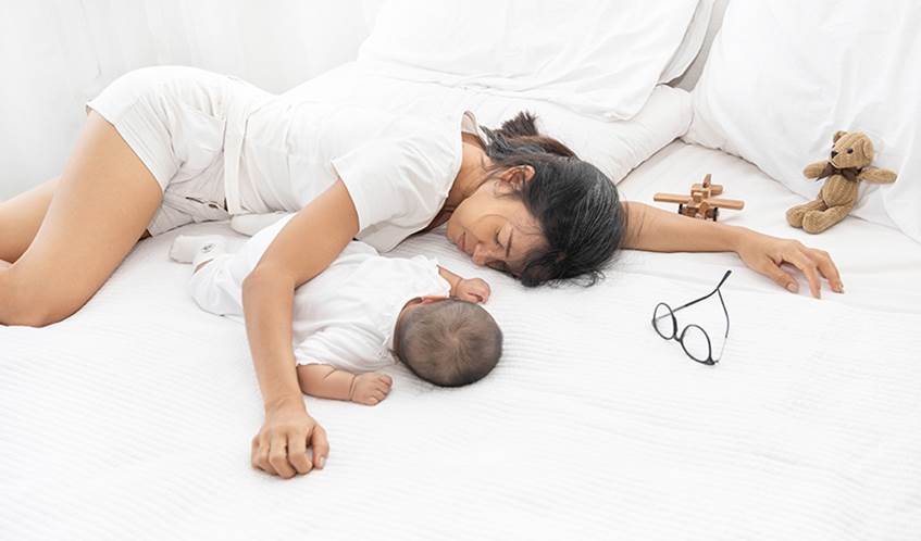 A mother lies on a bed alongside her young baby