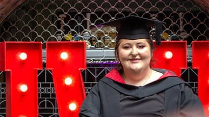 Close-up portrait of a graduate wearing a black gown with a red-lined hood and a black mortarboard. The graduate is seated in front of large illuminated red letters from a "UWE" display, with decorative metalwork and an arched architectural feature in the background. Bright bulb lights within the letters frame the graduate, creating a vivid contrast against the darker surroundings.