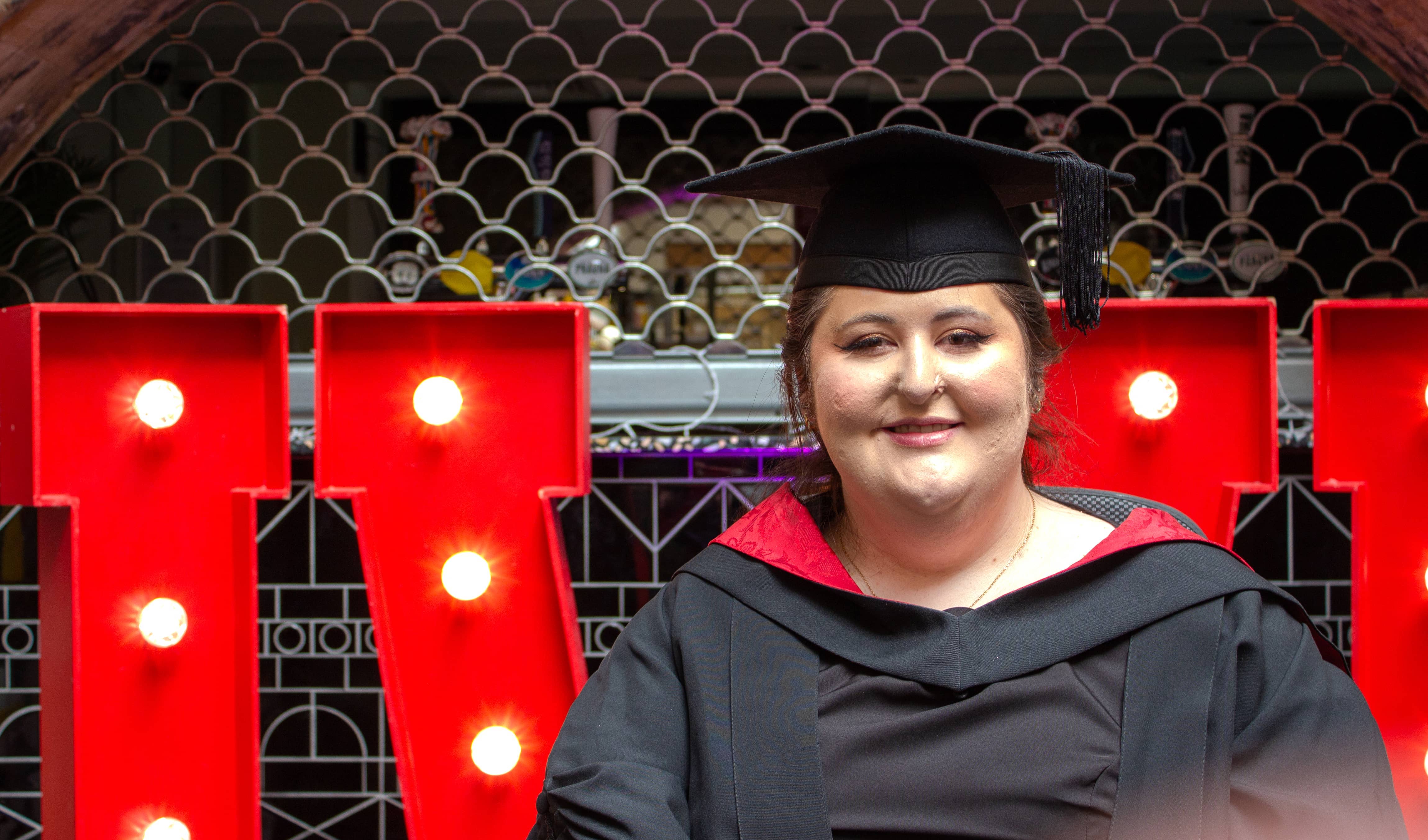 Close-up portrait of a graduate wearing a black gown with a red-lined hood and a black mortarboard. The graduate is seated in front of large illuminated red letters from a "UWE" display, with decorative metalwork and an arched architectural feature in the background. Bright bulb lights within the letters frame the graduate, creating a vivid contrast against the darker surroundings.