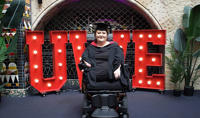 A graduate wearing a black gown with a red-lined hood and a black mortarboard sits in a powered wheelchair in front of large illuminated red letters spelling “UWE.” The scene is framed by a stone archway with decorative metalwork behind it, patterned tiled walls on the left, and tall green plants on either side. The glowing letters create a strong contrast against the darker interior background, with the graduate positioned centrally in the image.