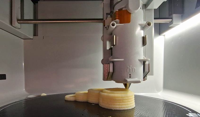 The inside of a 3D food printer, showing the machine produce a potato-based meal shaped in hoops
