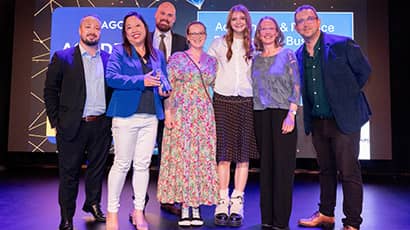 Seven people in a group smiling at the camera at an awards ceremony, with the word 'winner' displayed on a screen above their heads
