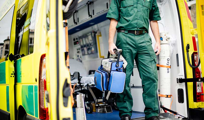Male paramedic standing in the back of an open ambulance, carrying a bag of kit in one hand