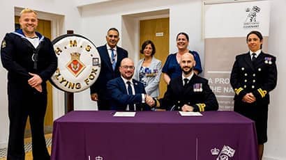 Professor Marc Griffiths (from UWE Bristol) and Lieutenant Commander Dominic Simonis-Law seated at a formal signing ceremony, with five other people stood behind them