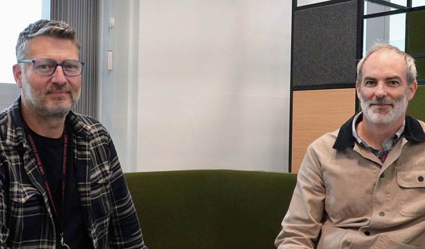 Two academics seated on a green sofa looking into the camera