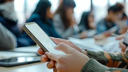Hands on a smartphone in the foreground, with blurred out people seated at desks in the background