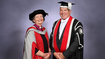 A woman wearing a graduation cap and gown smiles looking directly at the camera