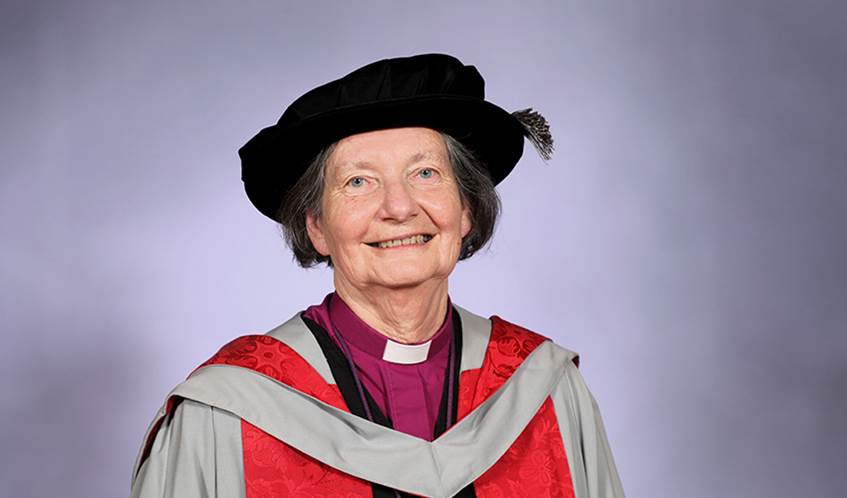 A woman wearing a graduation cap and gown smiles looking directly at the camera