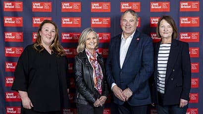 Three women and a man standing and smiling, posing for the camera in front of a branded red background