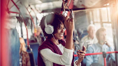 Lady wearing white over-ear headphones holding hand loop while standing on a busy bus.