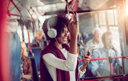 Lady wearing white over-ear headphones holding hand loop while standing on a busy bus.