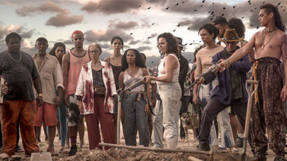 Cinematic scene of group of 15 people stood around earth mound, some holding pistols.