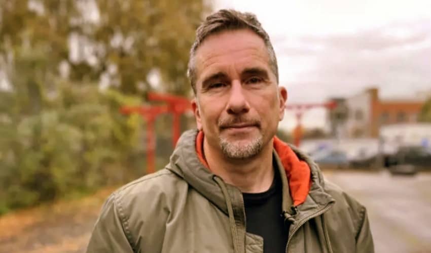 A man wearing a green coat looking at the camera, with a blurred background of buildings and vegetation