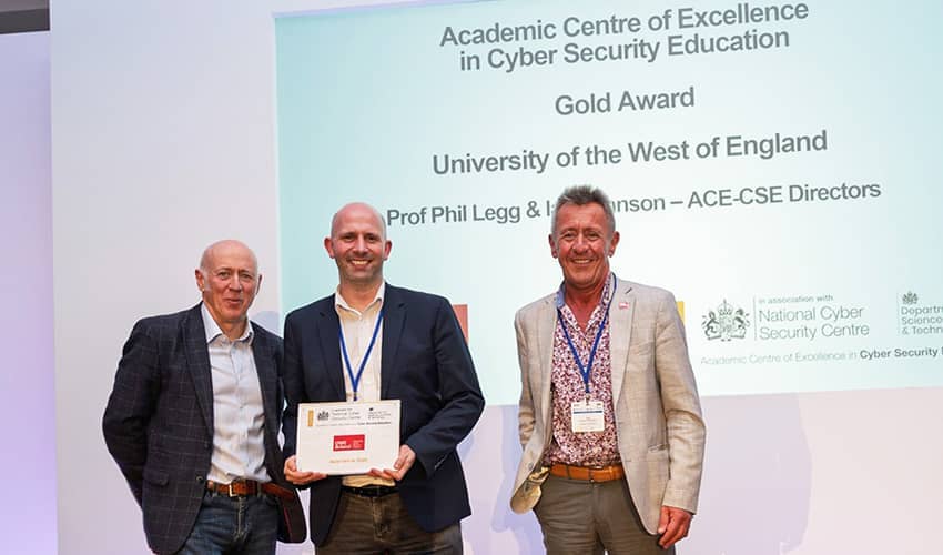 Three men wearing suits standing on a stage in front of a large screen smiling at the camera. The man in the centre of the group is holding a certificate.