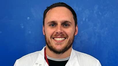 A bearded man wearing a white lab coat smiling at the camera, with a blue background