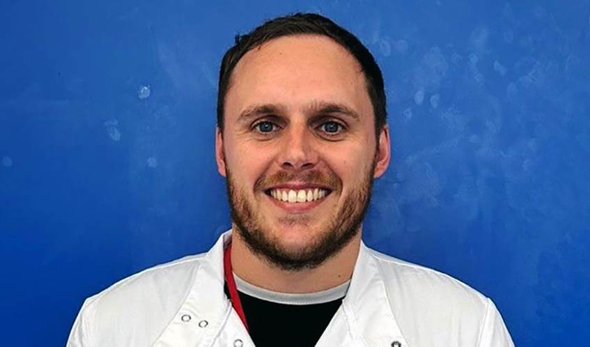 A bearded man in a white lab coat smiling at the camera, with a blue background