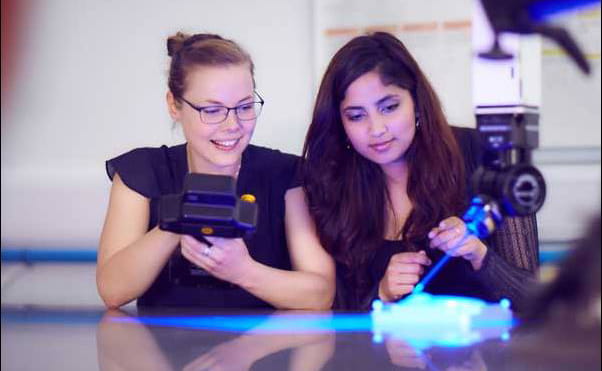 Two students holding equipment while blue light from machine shines onto desk in front of them