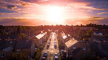 An urban area, focused on a street with terraced houses and parked cars, with the sun shining brightly in the background