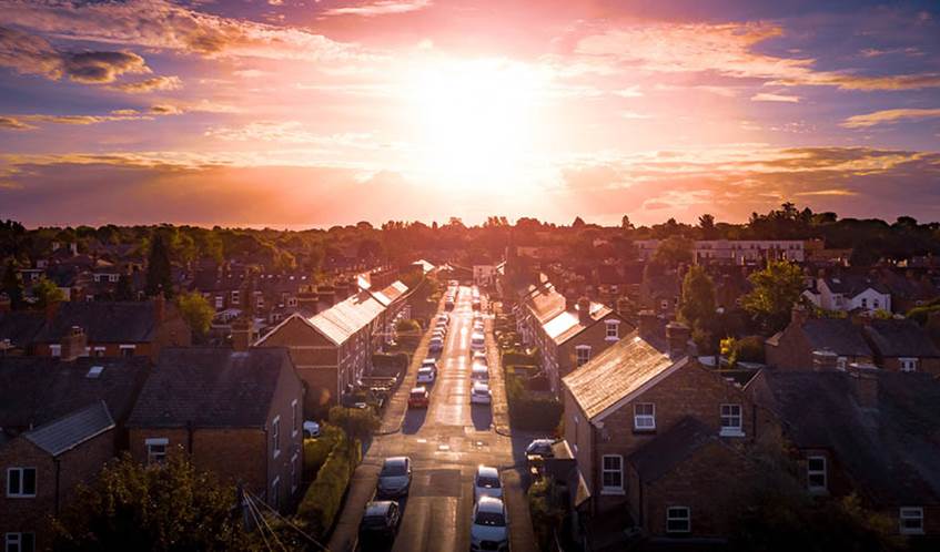 An urban area, focused on a street with terraced houses and parked cars, with the sun shining brightly in the background