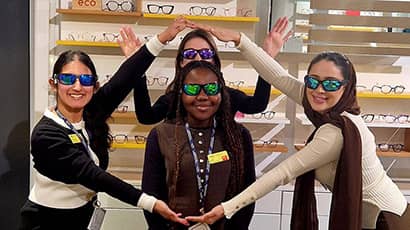 Four female students wearing sunglasses looking into the camera and smiling, with shelves of spectacle frames in the background