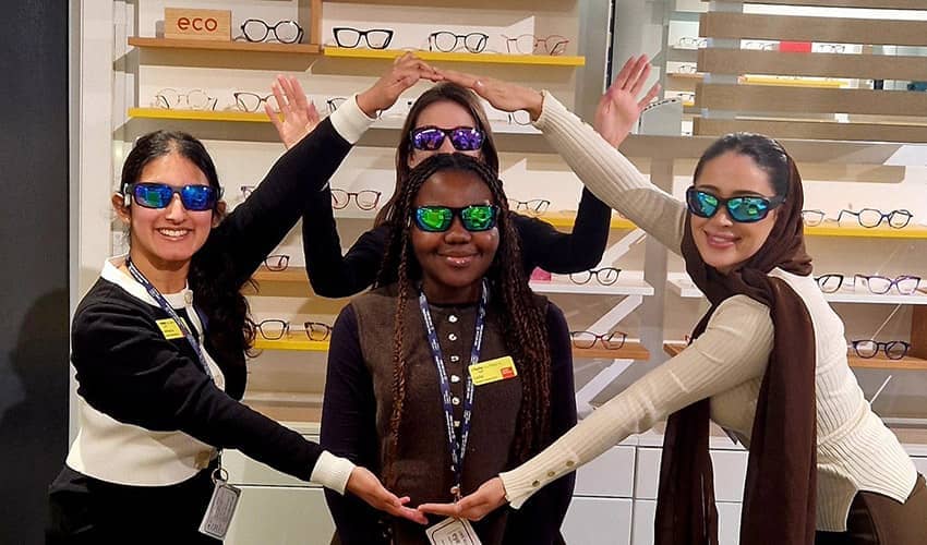 Four female students wearing sunglasses looking into the camera and smiling, with shelves of spectacle frames in the background