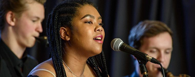 Woman singing into microphone with brass section in background.
