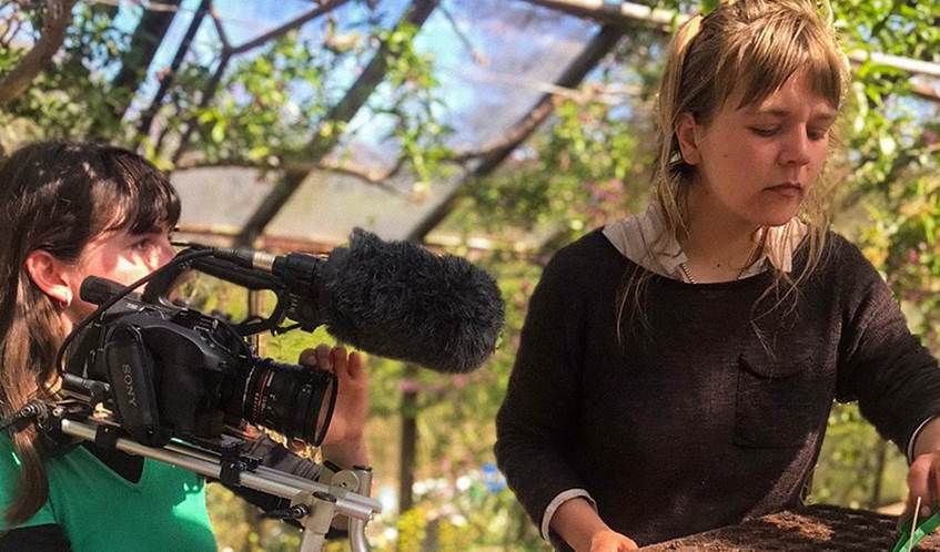 Two women in a greenhouse. One woman holds a film camera, filming another woman planting seeds in a tray.