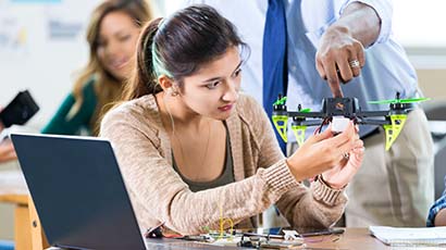 Female school student holds a drone she is building as her male teacher shows her something she needs to work on. A laptop and tools are on the table. Students are in the background working on projects.