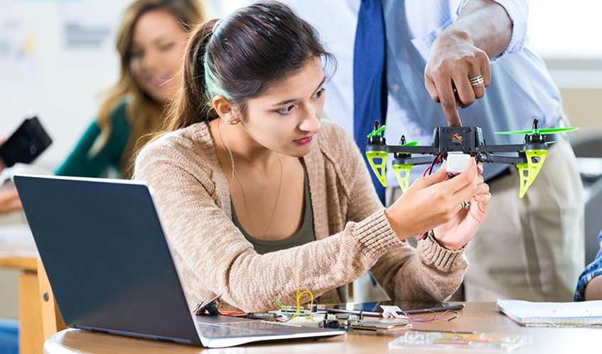 Female school student holds a drone she is building as her male teacher shows her something she needs to work on. A laptop and tools are on the table. Students are in the background working on projects.