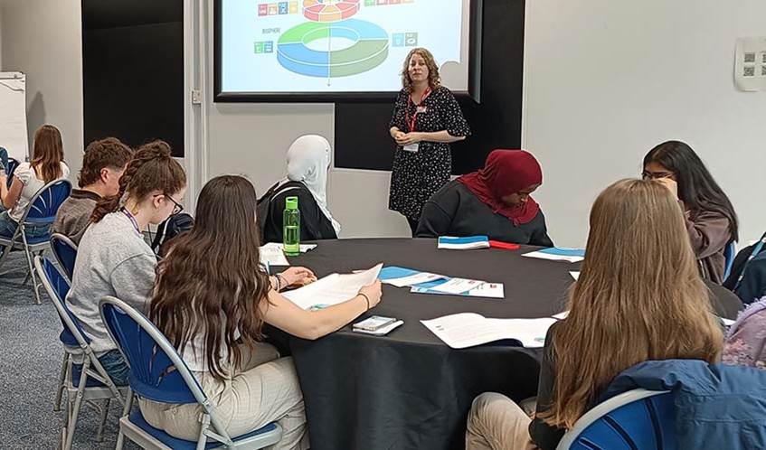 A classroom, with a lecturer stood in front of a white digital screen in the background, and secondary school students sat at a round table in the foreground
