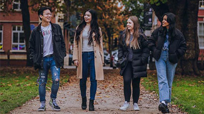 Group of four students walking along a campus path with autumn trees and leaves in background