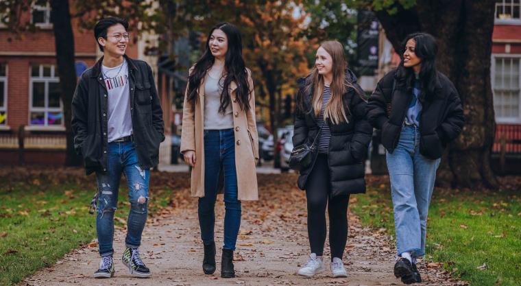 Group of four students walking along a campus path with autumn trees and leaves in background