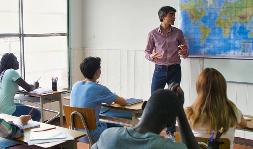 Image showing a man looking at a world map on the wall, teaching a group of secondary school children in a classroom