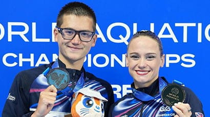 Two swimmers holding up their bronze medals and smiling after competing in a swimming event, with a blue background