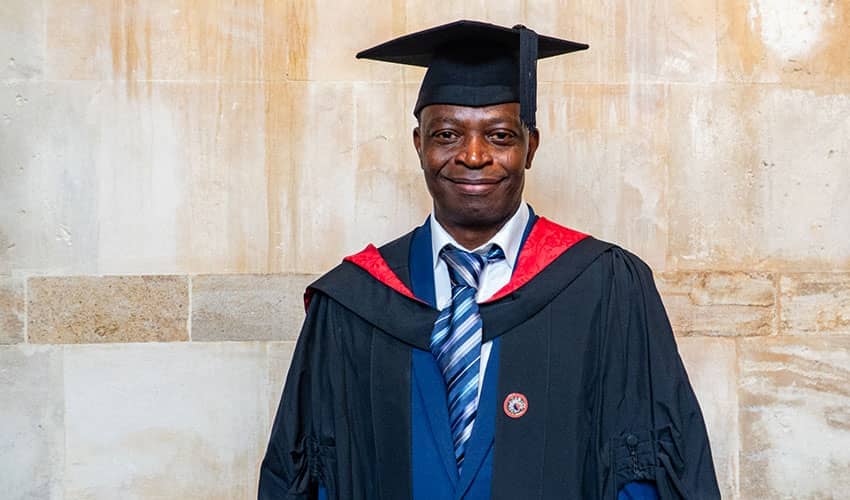 A man wearing graduation robes smiling at the camera, with a yellow stone background