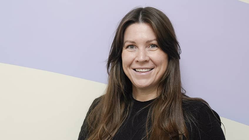 A person with long dark hair and wearing a dark jumper smiling at the camera, with a lilac and biege background