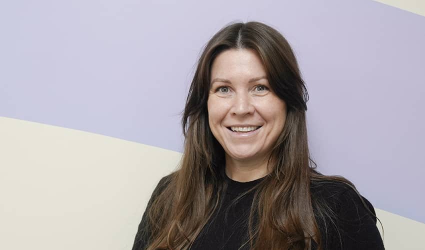 A person with long dark hair and wearing a dark jumper smiling at the camera, with a lilac and biege background