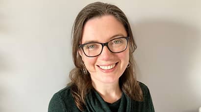 A woman wearing glasses smiles at the camera, with a grey background